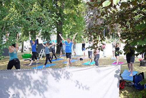 Hochschulsommerfest 20.06.18, Yoga, Foto: Stephanie Glöckner Im Bild befinden sich Teilnehmerinnnen und Teilnehmern im Stand mit nach oben gestreckten Armen in einer Yogastunde. Der Yogabereich wird im vorderen Bereich durch Tücher abgegrenzt.